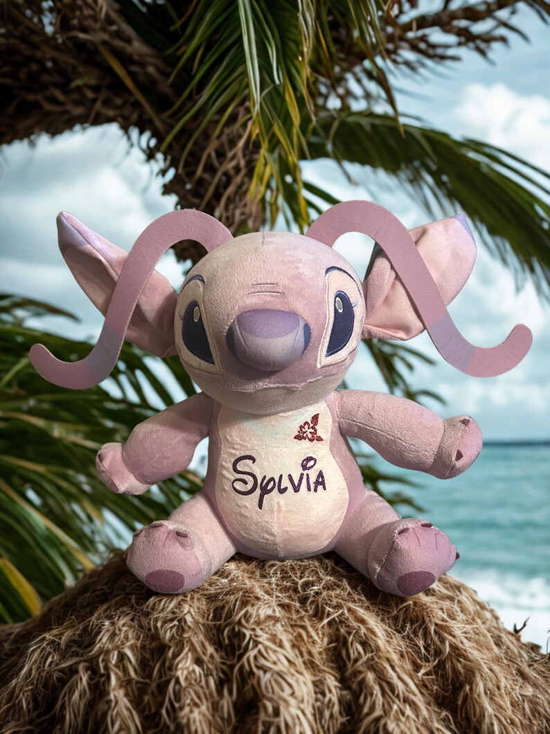 May include: A lavender-coloured plush toy with large ears and a smiling face. The word "Sylvia" is embroidered on the chest. The toy is sitting on a bed of straw, with a tropical background of palm leaves and ocean.
