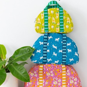 May include: Three colorful, patterned handbags stacked on top of each other. The top bag is yellow-green with a bunny print, the middle bag is blue with a white dog print, and the bottom bag is pink with a floral print. The text "Sunshine Sisters Handbag" is at the top.