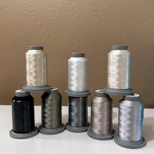 May include: Eight spools of thread in various colors, including black, gray, silver, and beige. The spools are arranged in two rows, with the top row having three spools and the bottom row having five spools.