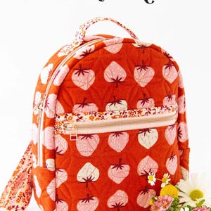 May include: A small orange backpack with a strawberry print. The backpack has a zippered pocket and a top handle. The text "THE VIOLET BACKPACK" is at the top of the image. The text "KNOT + THREAD design" is at the bottom of the image.
