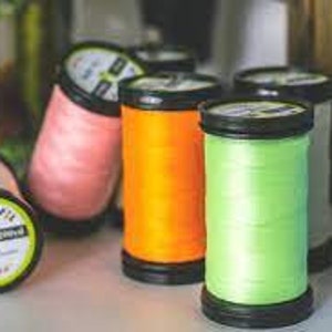 May include: A close-up of several spools of thread in various colors, including pink, orange, green, and blue. The spools are arranged in a row on a white surface.