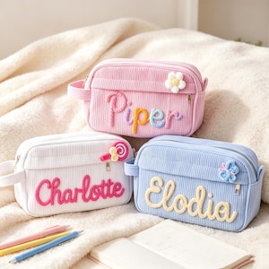 Può includere: Tre astucci rettangolari in velluto a coste nei colori rosa, bianco e azzurro. Ogni astuccio presenta una chiusura a zip, una maniglia laterale e un nome personalizzato in lettere di lana in rilievo: Piper, Charlotte ed Elodia. Le decorazioni in lana includono un fiore bianco, un lecca-lecca rosa e una farfalla blu.