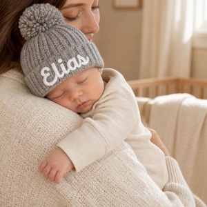 Può includere: Un berretto in maglia a coste grigio con un grande pompon sulla parte superiore. Il cappello presenta il nome Elias ricamato in corsivo bianco sul risvolto anteriore. Il berretto è realizzato in filato morbido e testurizzato con bordo ripiegato.