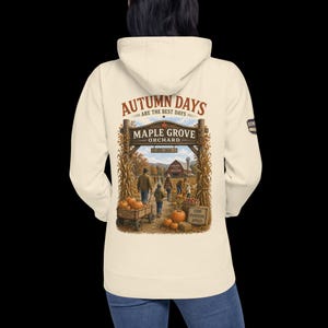 May include: A cream-colored hooded sweatshirt featuring a back graphic of an orchard entrance with the text "Autumn Days Are The Best Days" and "Maple Grove Orchard Est. 2020". The scene depicts people, a pumpkin wagon, and a red barn.