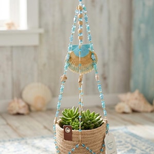 Può includere: Un portapiante sospeso con un cesto in corda intrecciata color marrone chiaro contenente una pianta succulenta verde. Il supporto è realizzato con perline blu e trasparenti, decorazioni a stella e una grande conchiglia dipinta di blu e marrone.