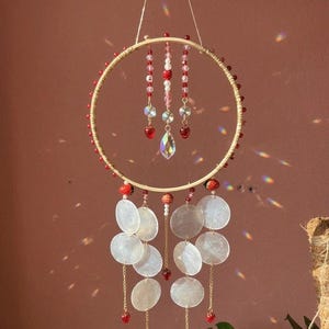 May include: A decorative dream catcher featuring a circular wooden frame embellished with red beads. Hanging from the top are strands of red, white, and clear beads, a large crystal, and heart-shaped charms. Below, white, iridescent shell-like discs dangle from the frame.