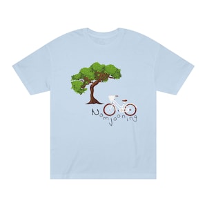 Puede incluir: Una camiseta azul claro con un gráfico de un árbol verde con tronco marrón y una bicicleta blanca con cesta delantera aparcada debajo. La palabra Namjooning está impresa en negro con un estilo de letra manuscrita bajo la bicicleta.