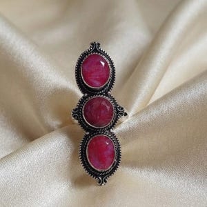May include: A silver ring featuring three vertically aligned oval-shaped red gemstones. Each stone is set within a decorative, twisted metal border. The design showcases a long, slender profile with intricate metalwork surrounding the stones.