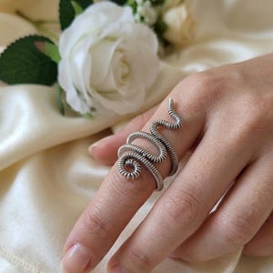 Może przedstawiać: Srebrzysty pierścionek o wzorze ciągłego, zwiniętego drutu, który owija się wokół palca w spiralny wzór. Metal ma teksturowany, żebrowany wygląd stworzony przez ciasno nawinięty drut, tworząc elastyczny i regulowany kształt pierścionka.