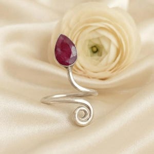 May include: A silver ring featuring a teardrop-shaped deep red gemstone at one end and a spiral coil design at the other. The band is made of a single piece of silver wire that curves to form the open-ended shape of the ring.