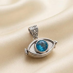 May include: A silver-toned pendant shaped like an eye, featuring a central round faceted blue gemstone. The metal frame has an oxidized finish with intricate scrollwork patterns on the bail and a smooth, polished border surrounding the stone.