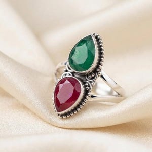 Puede incluir: Un anillo de plata con dos piedras preciosas facetadas en forma de pera dispuestas verticalmente. La piedra superior es de color verde intenso, mientras que la inferior es de un rojo intenso. El engaste metálico tiene una textura de cuentas.
