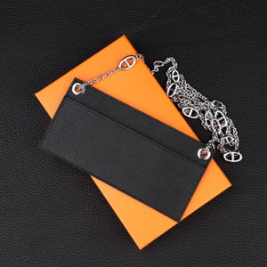 May include: A rectangular black leather card holder with a textured surface, featuring two silver-toned metal grommets at the top corners. A long silver-toned chain with oval-shaped links is attached to the grommets, resting on an orange box.