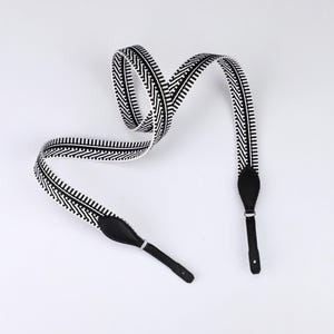 May include: A camera strap featuring a black and white woven pattern with a repeating chevron and striped design. The strap includes black leather end tabs with silver-toned metal hardware and a single punched hole at each end for attachment.