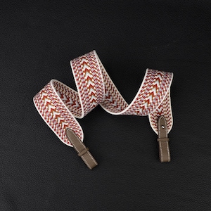 May include: A woven bag strap featuring a repeating chevron pattern in shades of red, white, and blue. The strap ends are finished with taupe-colored leather tabs, each featuring a single silver-toned metal stud and a small leather loop for attachment.