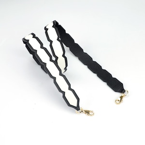 May include: A replacement bag strap featuring a black and white scalloped edge design. The strap is constructed with a textured black exterior and a smooth white interior, finished with gold-toned metal lobster claw clasps at both ends.