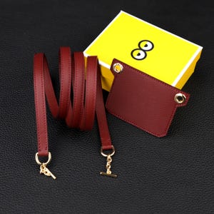 May include: A dark red leather strap with gold-toned metal hardware and a matching rectangular leather tag featuring two gold-toned eyelets. The set is displayed alongside a bright yellow box decorated with a graphic of two cartoon eyes.
