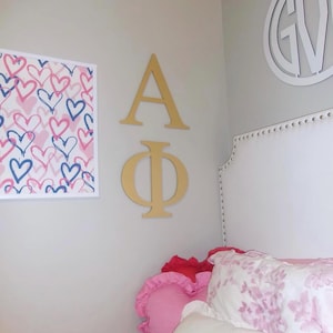 Op de afbeelding: Een slaapkamer met een ingelijste print met roze en blauwe harten, gouden Griekse letters en een wit hoofdbord. Het bed heeft roze en witte bloemenkussens en een hartvormig kussen.
