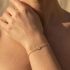 Op de afbeelding: Een delicate goudkleurige kettingarmband met een kleine, platte, zilverkleurige spookhanger met twee uitgesneden oogjes. De hanger is gecentreerd op de dunne metalen schakelketting die met een eenvoudige sluiting om de pols wordt bevestigd.