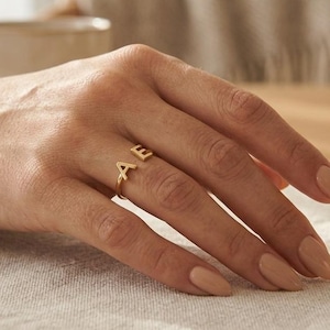 Peut inclure: Une bague ouverte dorée ornée de deux lettres majuscules, A et E, situées aux extrémités de l'anneau. La bague présente une finition métallique polie et un design simple et épuré qui repose sur le doigt, mettant les lettres en évidence.
