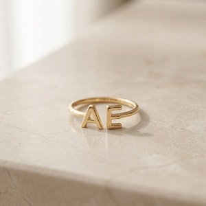 May include: A gold-toned open ring featuring two capital letters, A and E, positioned at the ends of the band. The ring has a polished, smooth finish and a simple, thin circular band design that remains open between the two distinct letter characters.