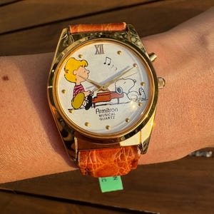 Puede incluir: Un reloj de pulsera Armitron Musical Quartz con caja de metal en tono dorado y correa de cuero naranja texturizada. La esfera blanca presenta un gr&aacute;fico de Schroeder tocando el piano con Snoopy, manecillas doradas y el texto Armitron Musical Quartz.