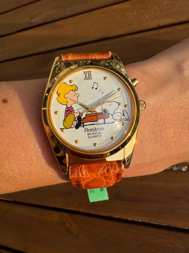 Puede incluir: Un reloj de pulsera Armitron Musical Quartz con caja de metal en tono dorado y correa de cuero naranja texturizada. La esfera blanca presenta un gr&aacute;fico de Schroeder tocando el piano con Snoopy, manecillas doradas y el texto Armitron Musical Quartz.