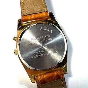Puede incluir: Un reloj de pulsera dorado con correa de cuero marr&oacute;n. La parte posterior de la caja de acero inoxidable presenta el texto ALIGNA, BASE METAL BEZEL, STAINLESS STEEL BACK, 2200/117, 400 101, THAILAND MOVT y TM & &copy; 1994 Warner Bros. grabado en el centro.