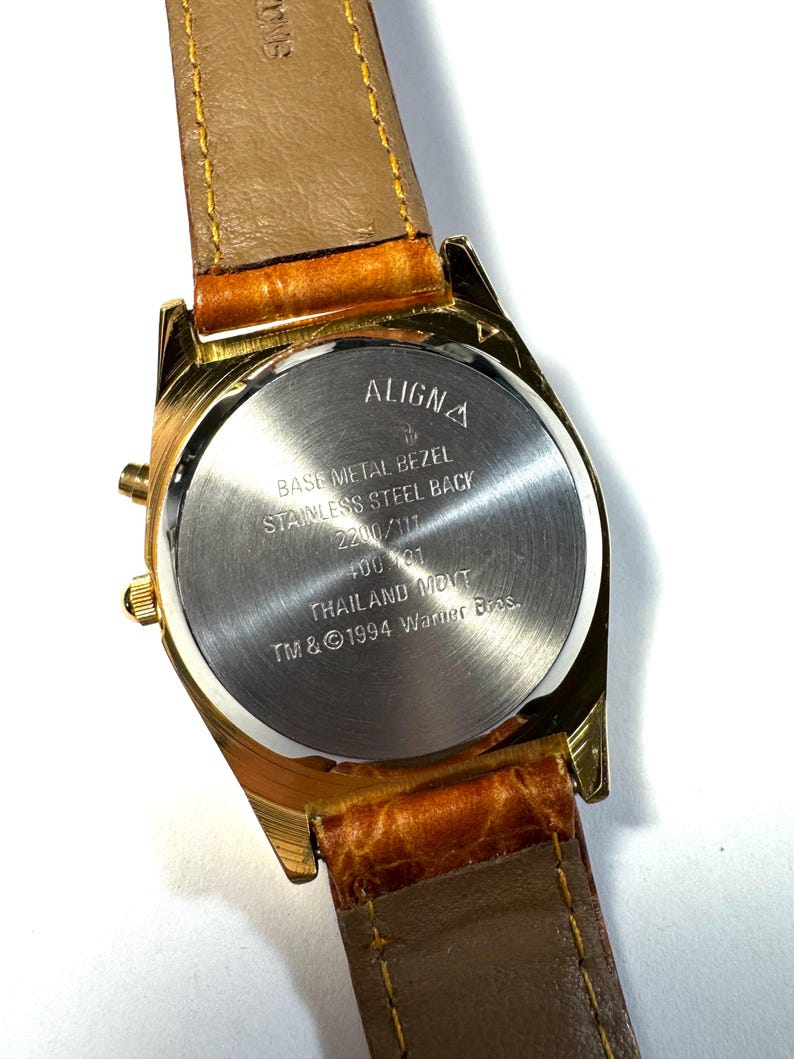 Puede incluir: Un reloj de pulsera dorado con correa de cuero marr&oacute;n. La parte posterior de la caja de acero inoxidable presenta el texto ALIGNA, BASE METAL BEZEL, STAINLESS STEEL BACK, 2200/117, 400 101, THAILAND MOVT y TM & &copy; 1994 Warner Bros. grabado en el centro.