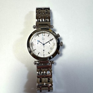May include: A silver-tone analog wristwatch with a round case and a matching link bracelet. The white textured dial features the Gc logo, three sub-dials, a date window at the four o'clock position, and blue-toned hands with Arabic numerals.