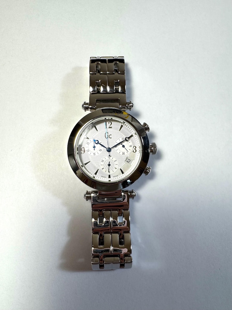May include: A silver-tone analog wristwatch with a round case and a matching link bracelet. The white textured dial features the Gc logo, three sub-dials, a date window at the four o'clock position, and blue-toned hands with Arabic numerals.