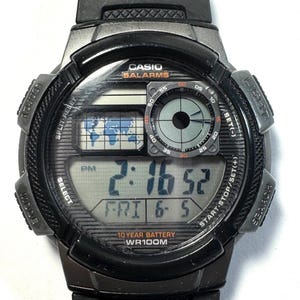 May include: A black Casio digital watch with a resin strap. The face features a world map display, a circular analog-style sub-dial, and a digital screen showing 2:16 PM, FRI 6-5. Text includes CASIO, 5ALARMS, 10 YEAR BATTERY, and WR100M.