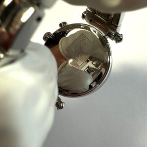 May include: A silver-toned wristwatch featuring a round case with a polished metallic back. The case back is engraved with the Gc logo and text, including the brand name and water resistance specifications, visible on the reflective surface.