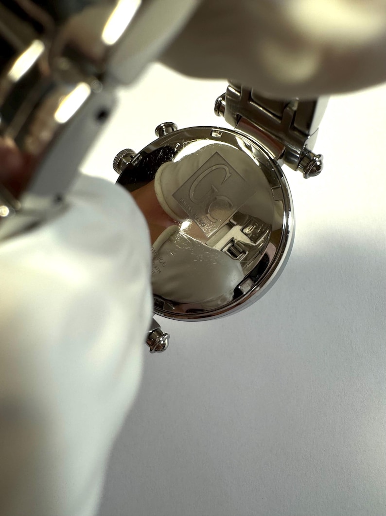 May include: A silver-toned wristwatch featuring a round case with a polished metallic back. The case back is engraved with the Gc logo and text, including the brand name and water resistance specifications, visible on the reflective surface.