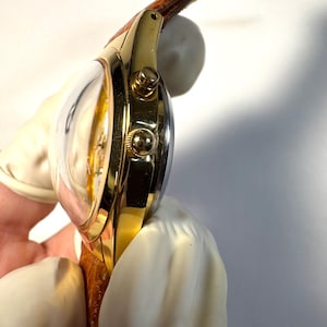 Puede incluir: Un reloj de pulsera dorado con un cristal abombado prominente, una caja pulida de color oro y dos botones laterales. El reloj cuenta con una correa de cuero marr&oacute;n texturizada unida a las asas, mostrando una vista lateral del reloj.
