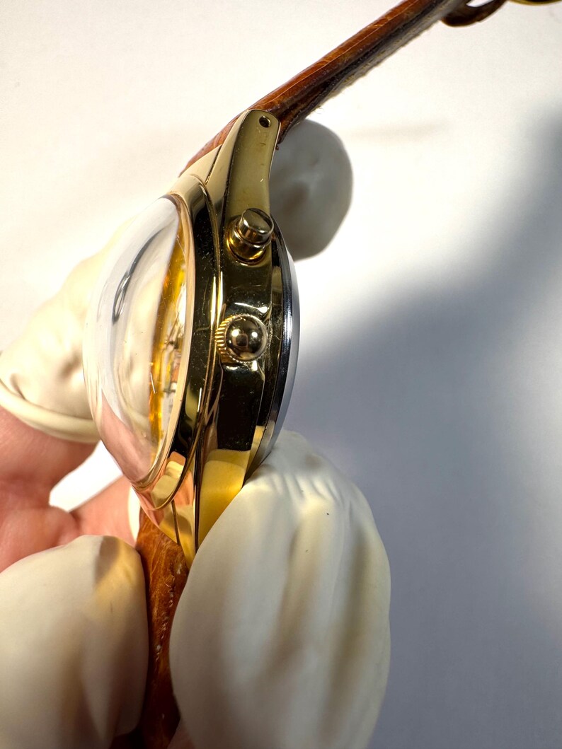 Puede incluir: Un reloj de pulsera dorado con un cristal abombado prominente, una caja pulida de color oro y dos botones laterales. El reloj cuenta con una correa de cuero marr&oacute;n texturizada unida a las asas, mostrando una vista lateral del reloj.