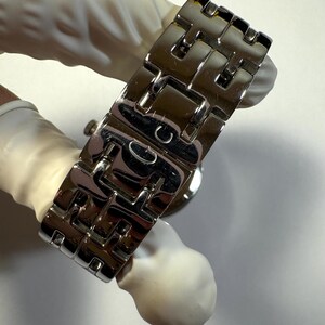 May include: A silver-toned metal watch bracelet featuring a series of interlocking H-shaped links. The polished surface reflects light, and the central clasp area displays a small engraved letter G, showcasing a structured and metallic design.