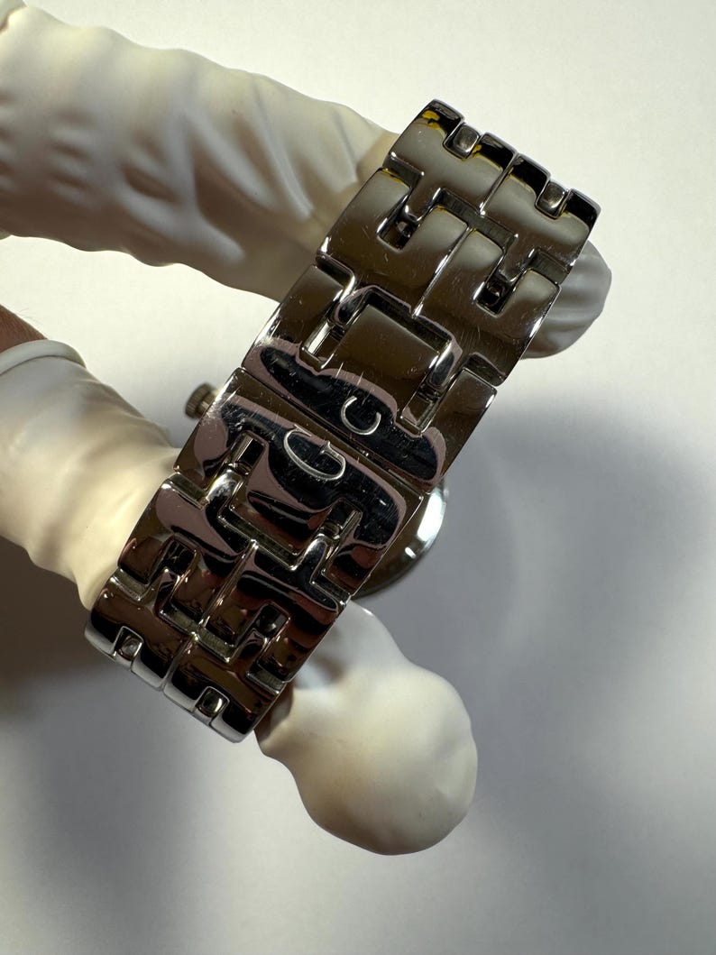 May include: A silver-toned metal watch bracelet featuring a series of interlocking H-shaped links. The polished surface reflects light, and the central clasp area displays a small engraved letter G, showcasing a structured and metallic design.