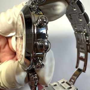 May include: A polished silver-tone stainless steel chronograph wristwatch featuring a round case, a matching link bracelet with a deployment clasp, and three side-mounted pushers and a central crown on the right side of the watch casing.