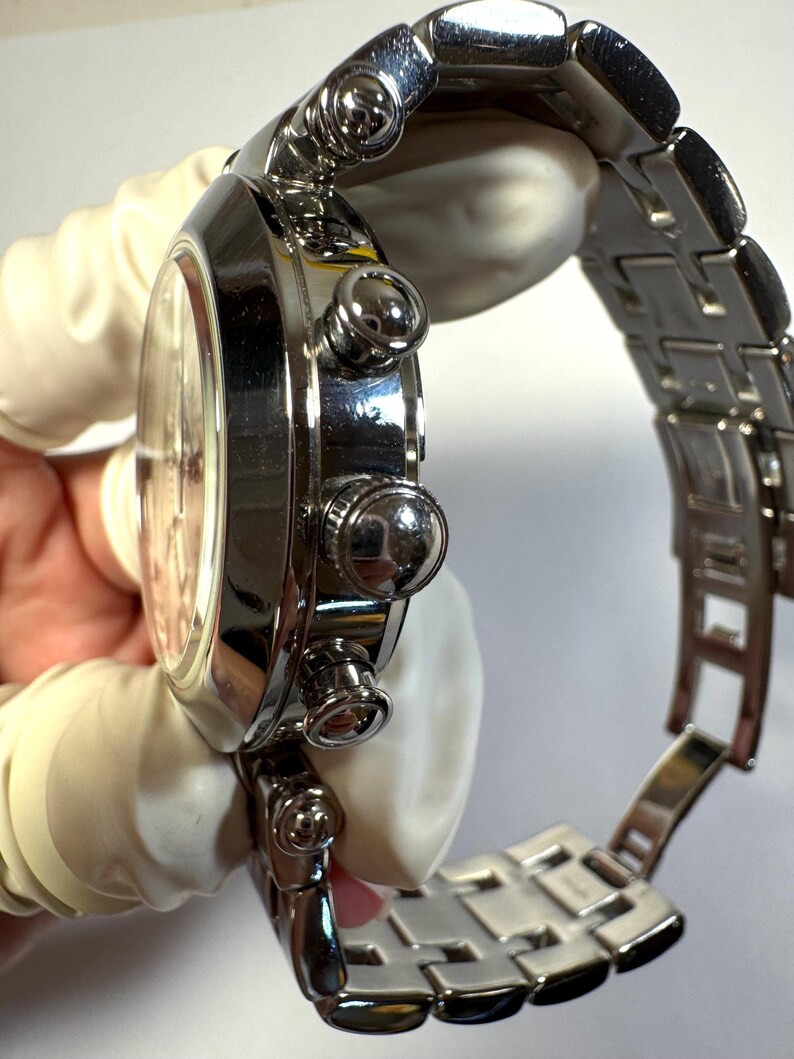 May include: A polished silver-tone stainless steel chronograph wristwatch featuring a round case, a matching link bracelet with a deployment clasp, and three side-mounted pushers and a central crown on the right side of the watch casing.
