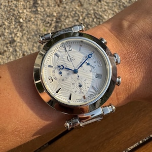 May include: A silver-tone chronograph wristwatch featuring a textured white dial with blue hands, three sub-dials, and the Gc logo at the twelve o'clock position. The watch includes a date window at the three o'clock position and Arabic numerals.