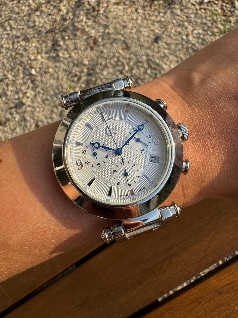 May include: A silver-tone chronograph wristwatch featuring a textured white dial with blue hands, three sub-dials, and the Gc logo at the twelve o'clock position. The watch includes a date window at the three o'clock position and Arabic numerals.