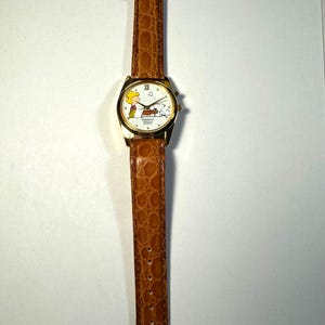 Puede incluir: Un reloj de pulsera dorado con una correa de piel sint&eacute;tica marr&oacute;n con textura de caim&aacute;n. La esfera blanca presenta un gr&aacute;fico de Schroeder tocando el piano con Snoopy, el n&uacute;mero romano XII arriba y el texto Remtron Musical Quartz.