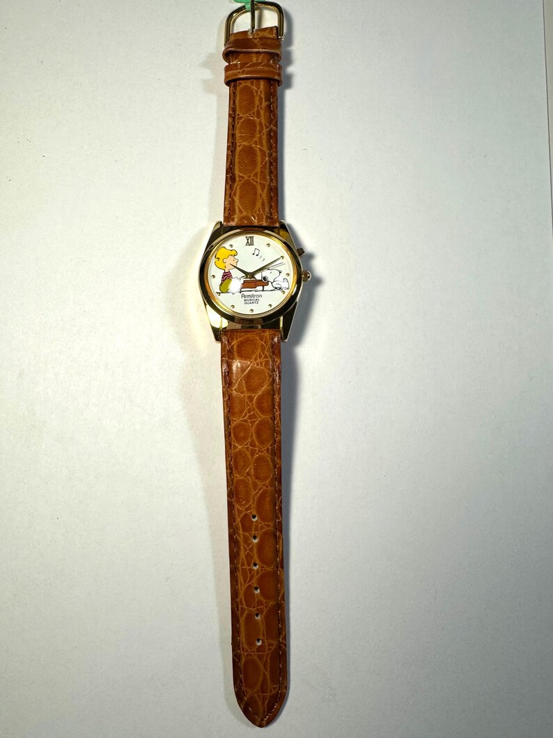Puede incluir: Un reloj de pulsera dorado con una correa de piel sint&eacute;tica marr&oacute;n con textura de caim&aacute;n. La esfera blanca presenta un gr&aacute;fico de Schroeder tocando el piano con Snoopy, el n&uacute;mero romano XII arriba y el texto Remtron Musical Quartz.