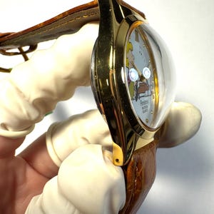 Puede incluir: Un reloj de pulsera dorado con cristal abombado, que presenta una esfera clara con una ilustraci&oacute;n de personaje de dibujos animados, notas musicales y el texto Armitron Musical Quartz. Tiene una correa de cuero marr&oacute;n texturizada.