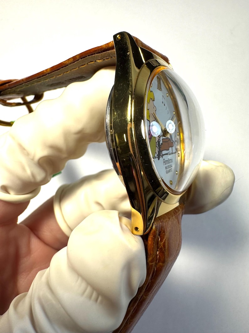 Puede incluir: Un reloj de pulsera dorado con cristal abombado, que presenta una esfera clara con una ilustraci&oacute;n de personaje de dibujos animados, notas musicales y el texto Armitron Musical Quartz. Tiene una correa de cuero marr&oacute;n texturizada.