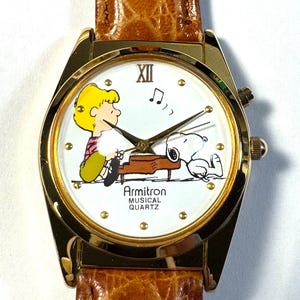 Puede incluir: Un reloj de pulsera Armitron Musical Quartz con caja dorada y correa de cuero marr&oacute;n texturizado. La esfera blanca presenta un gr&aacute;fico de Schroeder tocando el piano con Snoopy, manecillas doradas y el texto Armitron MUSICAL QUARTZ.