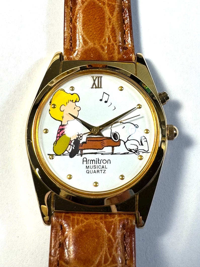 Puede incluir: Un reloj de pulsera Armitron Musical Quartz con caja dorada y correa de cuero marr&oacute;n texturizado. La esfera blanca presenta un gr&aacute;fico de Schroeder tocando el piano con Snoopy, manecillas doradas y el texto Armitron MUSICAL QUARTZ.