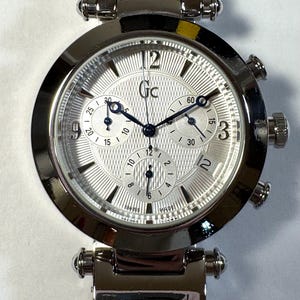 May include: A silver-tone stainless steel chronograph wristwatch with a round case and a textured silver dial. The watch features three sub-dials, a date window at the four o'clock position, dark blue hands, and the Gc logo printed on the dial face.