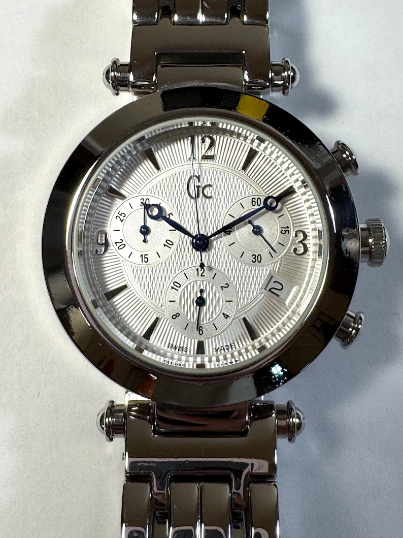 May include: A silver-tone stainless steel chronograph wristwatch with a round case and a textured silver dial. The watch features three sub-dials, a date window at the four o'clock position, dark blue hands, and the Gc logo printed on the dial face.